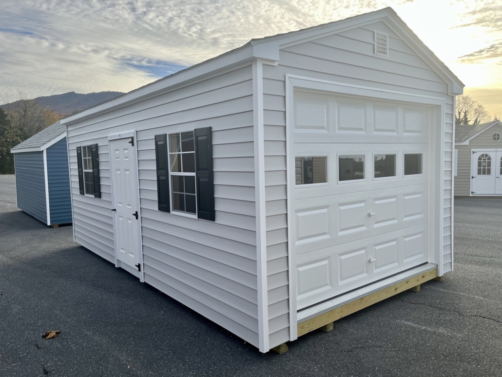 Garage with light siding, a white door and black shutters on pavement.