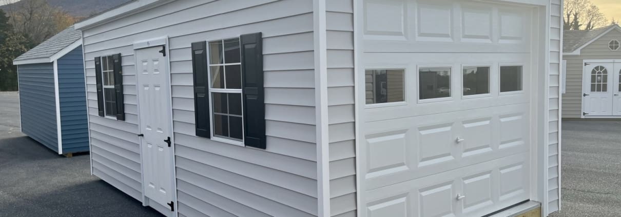 Garage with light siding, a white door and black shutters on pavement.