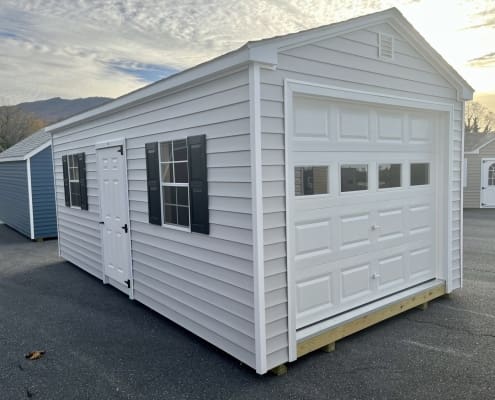 Garage with light siding, a white door and black shutters on pavement.